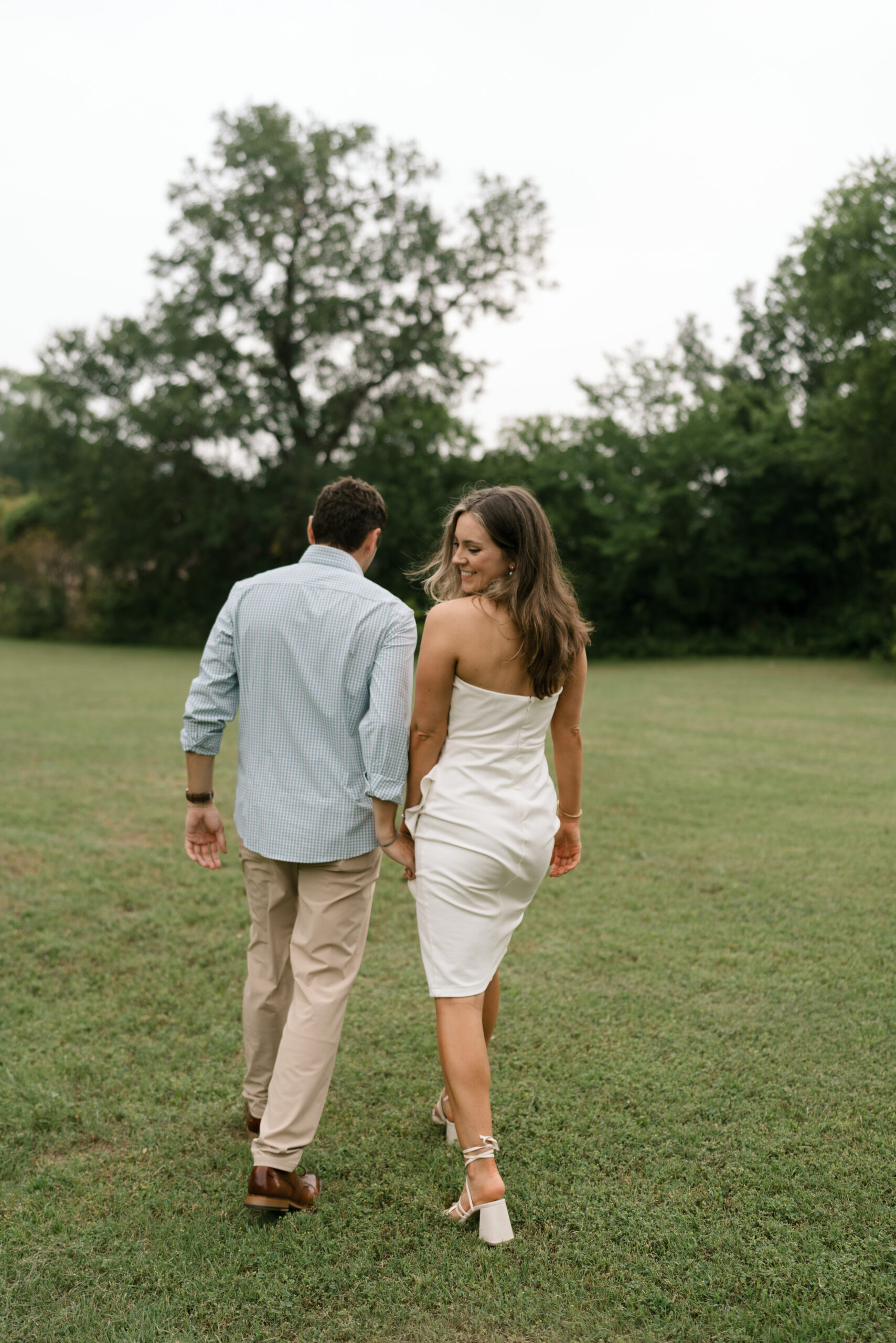 A couple, bride and groom, walk away from the camera during their rehearsal dinner. She is wearing a white dress and he is wearing. ablue shirt.
